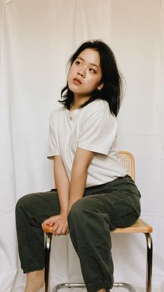 a woman sitting on top of a wooden chair next to a white wall and wearing green pants