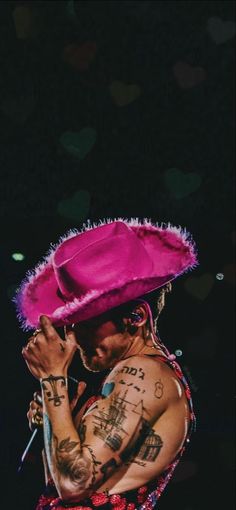 a tattooed man wearing a pink hat and holding his hands up to his face while standing in front of a black background