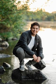 a man sitting on top of a rock next to a river with trees in the background