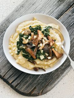 a white bowl filled with mashed potatoes covered in mushrooms and spinach on top of a wooden table