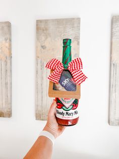 a hand holding a bottle of jam with a red and white bow on it in front of two paintings