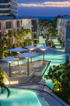 an outdoor swimming pool surrounded by palm trees and buildings at night with umbrellas over it