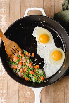 two fried eggs and vegetables in a skillet with a wooden spatula on the side