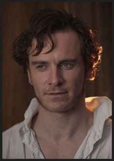 a man with curly hair and blue eyes looks at the camera while wearing a white shirt