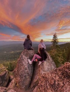 two people sitting on top of a large rock next to each other in front of a sunset