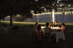 a group of people sitting around a table with candles in front of the water at night