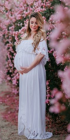 a pregnant woman wearing a white gown standing in front of pink flowers with her hands on her belly