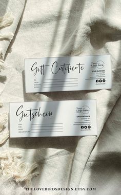 two gift certificates sitting on top of a white bed sheet with tasseled edges