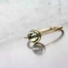 Olive Dot is a delicate yellow gold cabochon ring. A pale green sapphire from Montana is bezel set atop a round profile shank. Such a sweet design that can be stacked with other rings or worn by itself. I love the subtle color contrast between the olive green gemstone (5mm) and the warmth of the 14k yellow gold setting. If you love the idea of an understated American gemstone ring look no further! By the way, sapphire is September's birthstone. Shank measures 1.26mm. Gemstone weighs 0.81ct. Size Yellow Gold Domed Jewelry With Bezel Setting, Green Round Cut Birthstone Ring In 14k Gold, Green 14k Gold Round Cut Birthstone Ring, Elegant Green Cabochon Moonstone Ring, Classic Green Round Cut Sapphire Ring, Green Gemstone Ring Gold-plated, 14k Gold Cabochon Sapphire Ring, Green Sapphire Ring In 14k Gold, Round Cut, Montana Sapphire