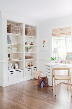 a home office with white bookcases and baskets on the desk, next to a window