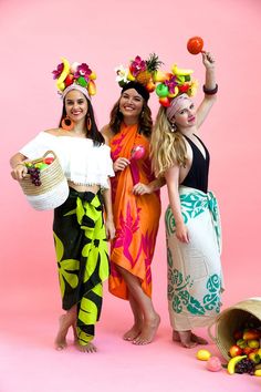 three beautiful women standing next to each other in front of a pink background with fruit on their heads