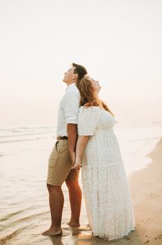 young newly engaged couple wearing white & beige hold hands, standing back to back while standing barefoot on the edge of the water St. Andrews State park, PCB Florida.  Portrait captured by Brittney Stanley of Be Seen Photos