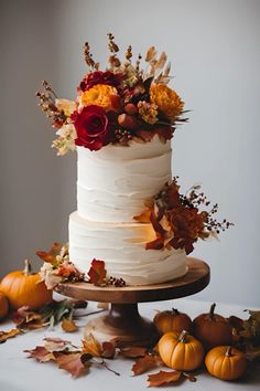 a three tiered white cake with flowers and leaves on the table next to pumpkins