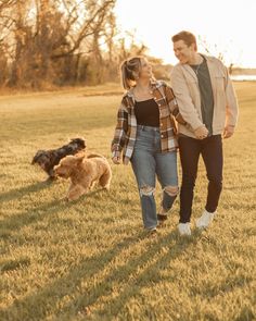 a man and woman walking with two dogs in a field at sunset, one has his hand on the dog's leash