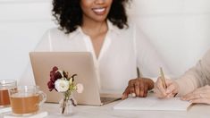 two women sitting at a table writing on paper with a laptop in front of them