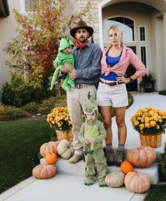 three people standing in front of a house with pumpkins on the ground and one person holding a stuffed animal