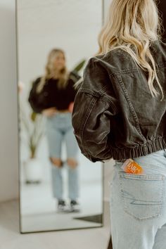 a woman standing in front of a mirror with her back to the camera, wearing jeans and a black jacket