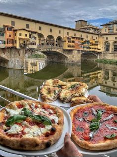 two pizzas are on plates in front of a river and bridge with buildings in the background