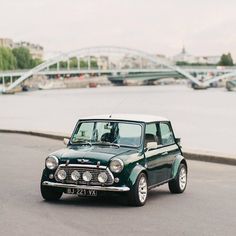 an old green car driving down the road next to a body of water with a bridge in the background