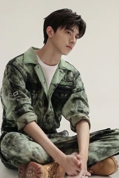 a young man sitting on the ground with his hands in his pockets and wearing camouflage clothing