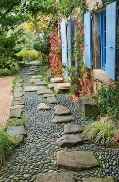 a stone path with blue shutters leading to a house in the distance, surrounded by greenery and flowers