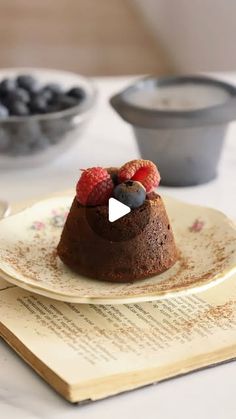 a chocolate cake with berries and blueberries on top sits on a book next to bowls of fruit