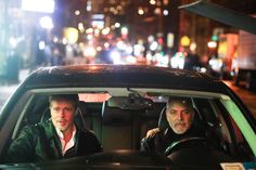 two men sitting in the driver's seat of a car on a city street at night