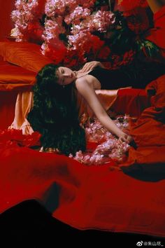 a woman laying on top of a bed covered in red sheets next to pink flowers