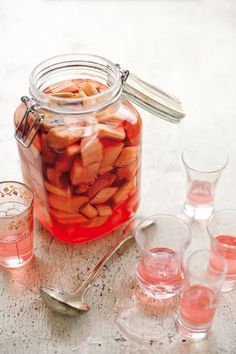 a jar filled with lots of red liquid next to glasses and spoons on top of a table