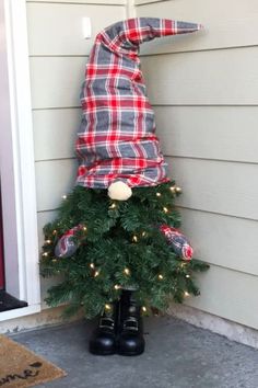 a christmas tree with a plaid hat on it's head sitting in front of a house