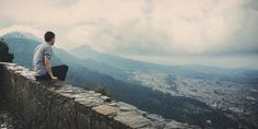 a man sitting on top of a stone wall next to a lush green hillside under a cloudy sky