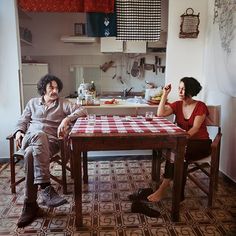 a man and woman sitting at a table in a kitchen with red checkered table cloth