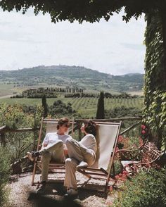 two people sitting on a bench in the middle of an outdoor area with flowers and greenery