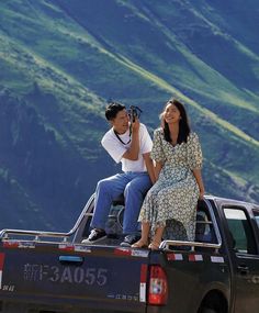 a man and woman sitting on the back of a pick up truck with mountains in the background