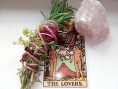 the lovers tarot card surrounded by flowers and herbs on a white surface with a pink rock