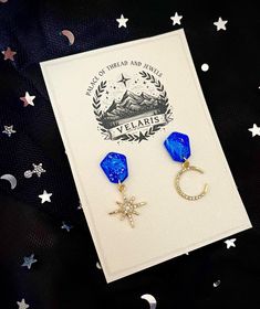 Celestial moon and star earring set with a stunning marbled blue stud that evokes the dark, seductive beauty of the Night Court.  The earrings are packaged like they were from Velaris' own Palace of Thread and Jewels, as if Rhysand himself personally picked it out.  This is the perfect ACOTAR gift to give.. or keep for yourself.  A base of translucent and star-speckled clay gives the stud an ethereal glow, and they are sealed with a shiny resin finish.    These are handmade polymer clay earrings. Colors and design will vary slightly from those pictured.  Celestial Stud Earrings, Mismatched Moon and Stars, ACOTAR Jewelry Gift, Night Court Earrings, Rhysand Gift, Velaris Book Jewelry, BookTok Acotar Jewelry, Bookish Crafts, The Night Court, Star Earring, Speckled Clay, Moon And Star Earrings, Night Court, Book Jewelry, Moon And Stars