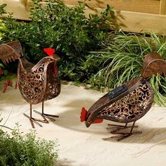 two metal roosters standing next to each other in front of some plants and flowers