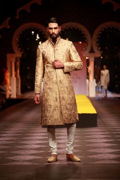 a man standing on a runway wearing a gold sherwanit and white pants