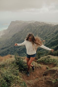 a woman is running up a hill with her hair blowing in the wind and mountains behind her