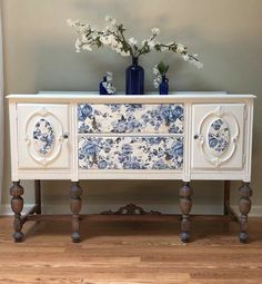 an ornate white and blue dresser with flowers on the top, in front of a gray wall