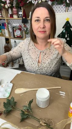 a woman sitting at a table with some crafting supplies on the table and christmas trees in the background