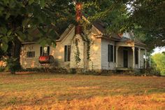 an old white house sitting in the middle of a field with lots of trees around it