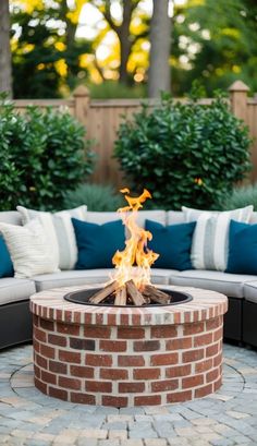 an outdoor fire pit with pillows on the side and trees in the back ground behind it