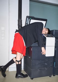 a man in black jacket and red skirt leaning over a printer machine with his head on it