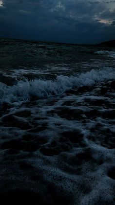 the sky is dark and cloudy at night over the ocean with waves crashing on the shore