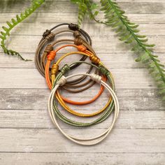 six bracelets with different colors and designs on wooden table next to fern leaves, top view