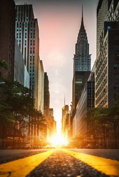 the sun is setting on a city street with tall buildings in the background and yellow lines painted on the road