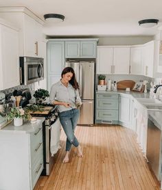 a woman standing in the middle of a kitchen with wooden floors and white cupboards