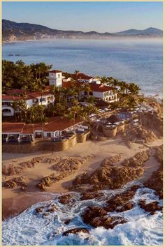 Aerial view of a coastal resort with white buildings and terracotta roofs overlooking a rocky shoreline with breaking waves, nestled against a backdrop of a town and distant hills. Romantic Escapes, Baja California Sur