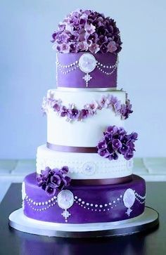 a three tiered cake with purple flowers and pearls on the top, sitting on a table